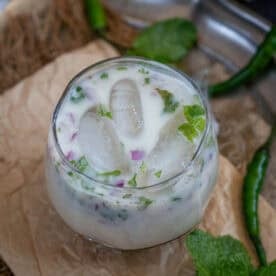 A refreshing glass of Sattu Chaas with ice cubes, chopped herbs, and red onion bits, garnished with mint leaves. Green chilies and mint leaves are visible in the background, making this traditional Sattu Chaas Recipe perfect for a cool summer drink.