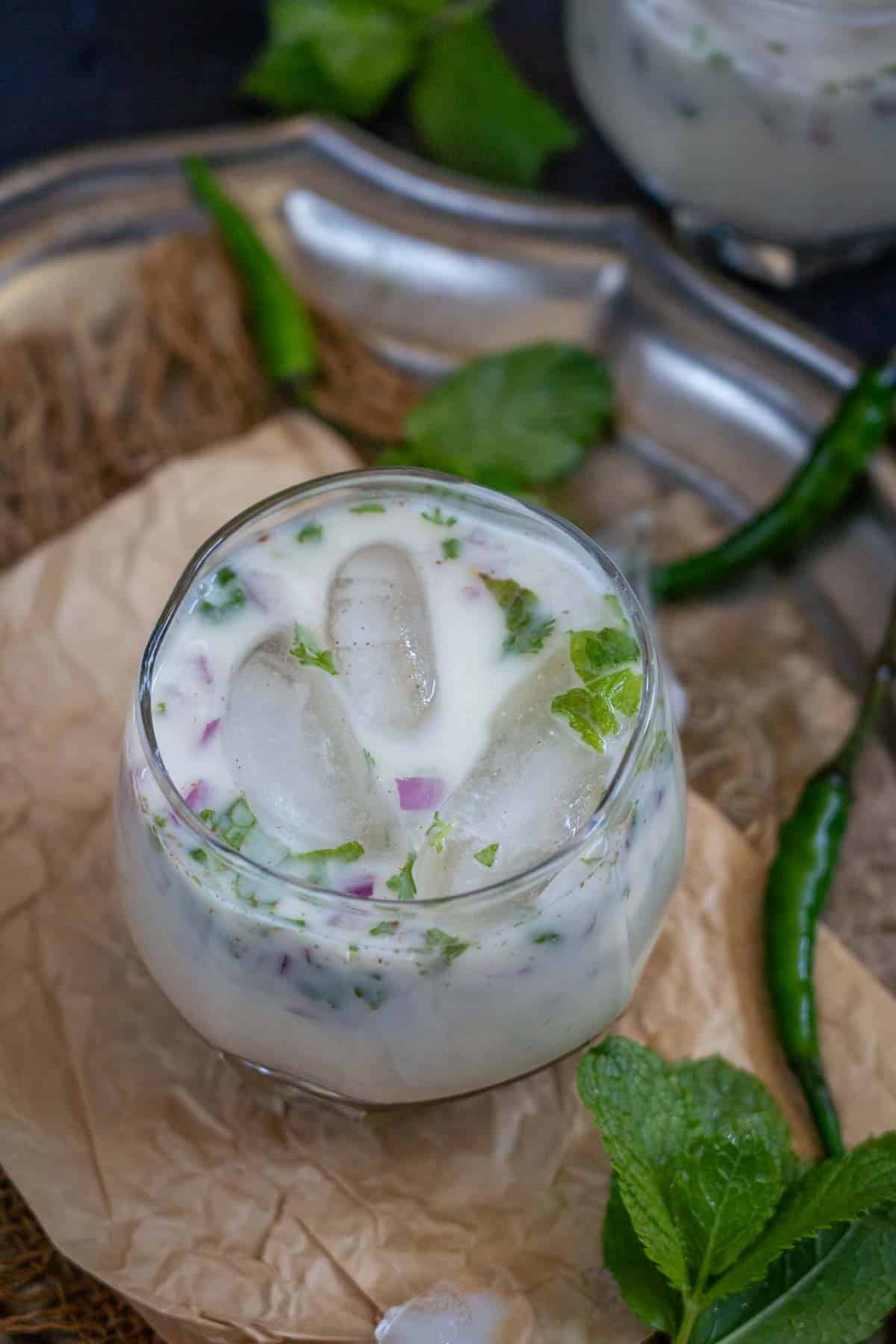 A refreshing glass of Sattu Chaas with ice cubes, chopped herbs, and red onion bits, garnished with mint leaves. Green chilies and mint leaves are visible in the background, making this traditional Sattu Chaas Recipe perfect for a cool summer drink.