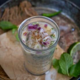 A glass filled with sattu drink topped with chopped red onions, herbs, and spices. Mint leaves and a lime wedge are visible in the blurred background.