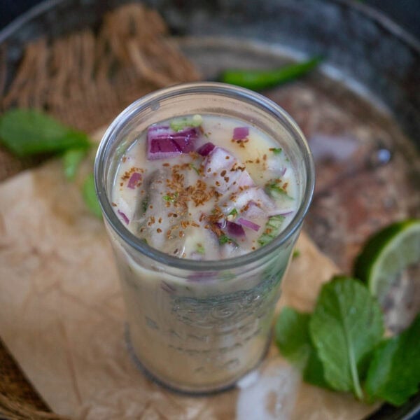 A glass filled with sattu drink topped with chopped red onions, herbs, and spices. Mint leaves and a lime wedge are visible in the blurred background.