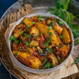 A bowl of spiced aloo baingan sabji garnished with fresh cilantro, placed on brown parchment paper with green herbs on the side.