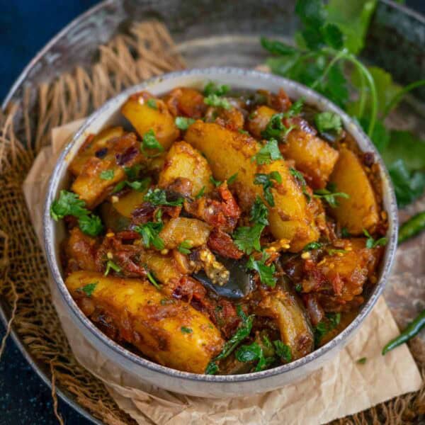 A bowl of spiced aloo baingan sabji garnished with fresh cilantro, placed on brown parchment paper with green herbs on the side.