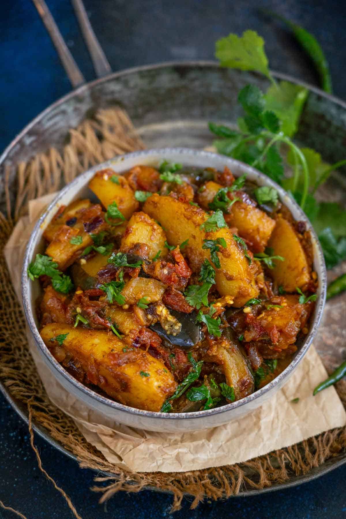A bowl of spiced aloo baingan sabji garnished with fresh cilantro, placed on brown parchment paper with green herbs on the side.