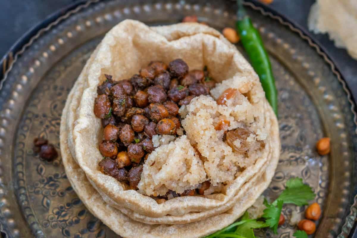 A metal plate with a stack of poori topped with sookha kala chana and a portion of sooji halwa, garnished with green chili and cilantro.