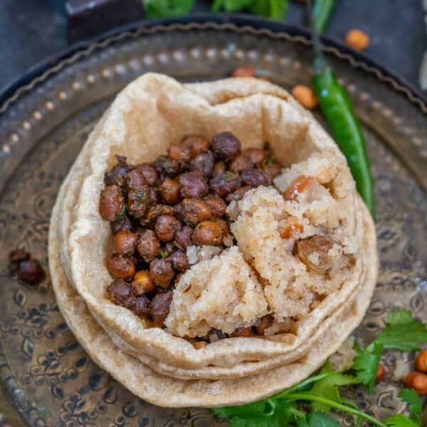 A metal plate with a stack of poori topped with sookha kala chana and a portion of sooji halwa, garnished with green chili and cilantro.