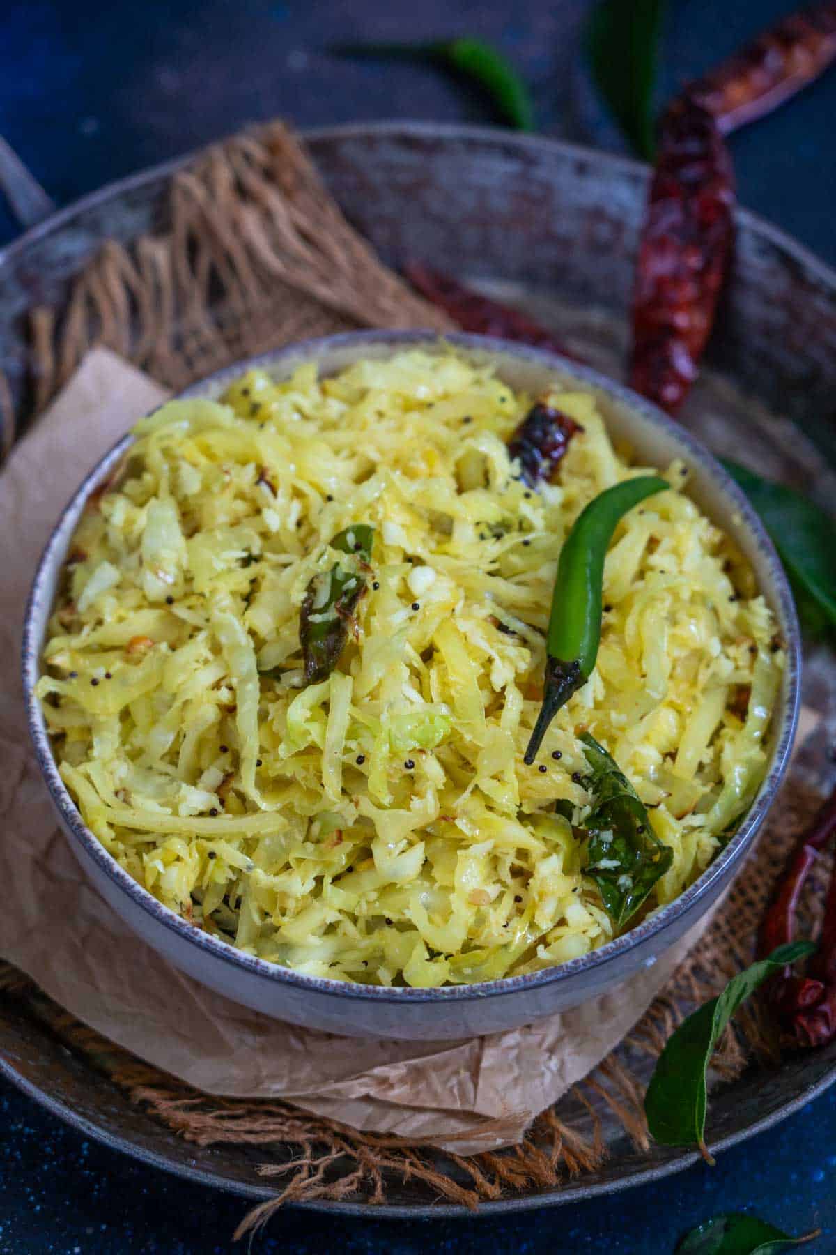 A bowl of cabbage poriyal stir-fry, garnished with mustard seeds, dried red chilies, curry leaves, and a green chili, served on a rustic surface—perfect for anyone searching for an authentic Cabbage Poriyal Recipe.