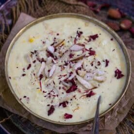 A bowl of creamy, lauki kheer garnished with sliced almonds, dried rose petals, and saffron strands, placed on a brown paper atop a dark rustic surface with a spoon on the side.