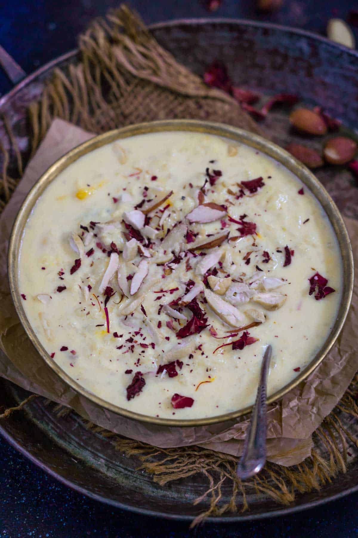 A bowl of creamy, lauki kheer garnished with sliced almonds, dried rose petals, and saffron strands, placed on a brown paper atop a dark rustic surface with a spoon on the side.